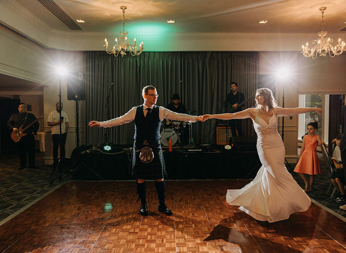 A bride in a wedding dress and a groom in a kilt dance together on a wooden dance floor in front of a small stage with a band