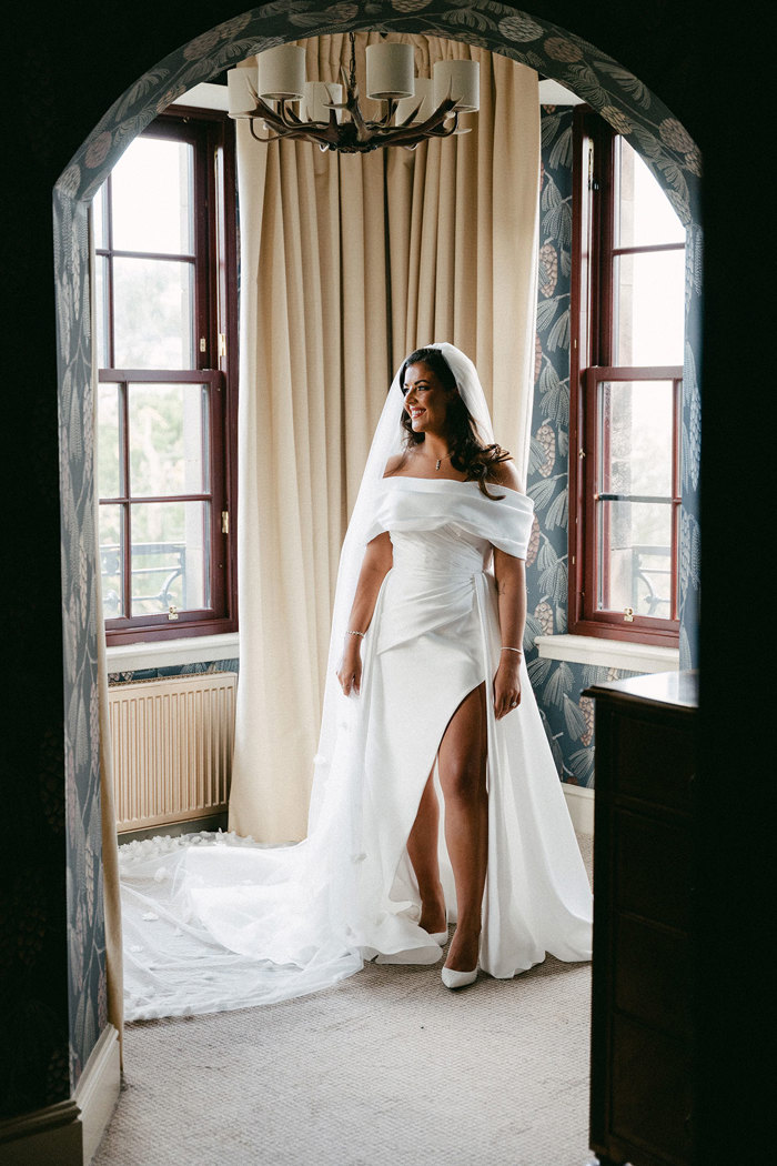 a brunette bride stands in the archway of a muted wallpapered room smiling and looking to one side as she wears a long trained wedding dress with off the shoulder cuff design and a high leg slit