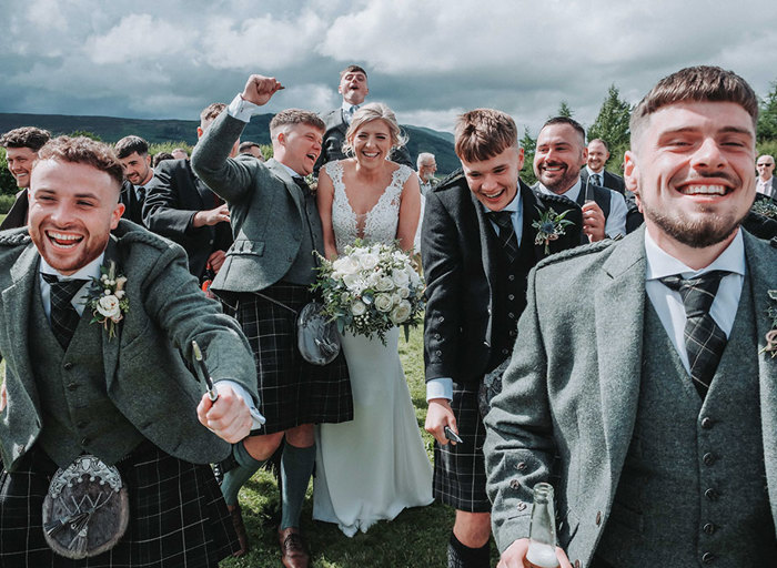 a bride and groom cheering and laughing as boisterous men in kilts run past them