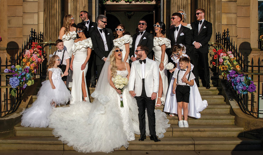 a wedding party posing and wearing sunglasses on a stone staircase outside a pillared doorway at Kimpton Blythswood Square Hotel in Glasgow. Rainbow coloured flowers adorn black railings either side of the stairs. 