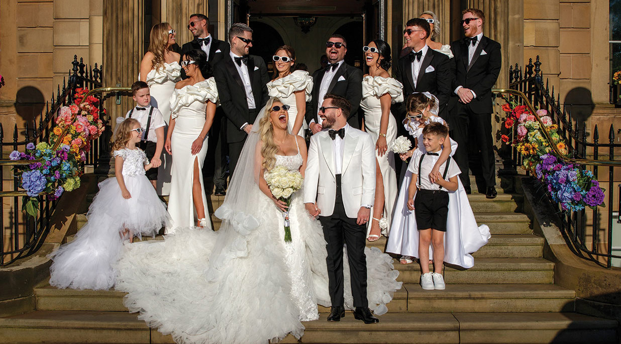 a wedding party posing and wearing sunglasses on a stone staircase outside a pillared doorway at Kimpton Blythswood Square Hotel in Glasgow. Rainbow coloured flowers adorn black railings either side of the stairs. 