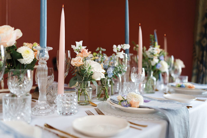 A table with a white tablecloth, pink and blue candles and pastel coloured flowers in small jars 