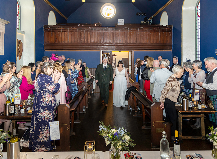 rows of wedding guests standing next to wooden pews clap as a bride and groom walk down the aisle.