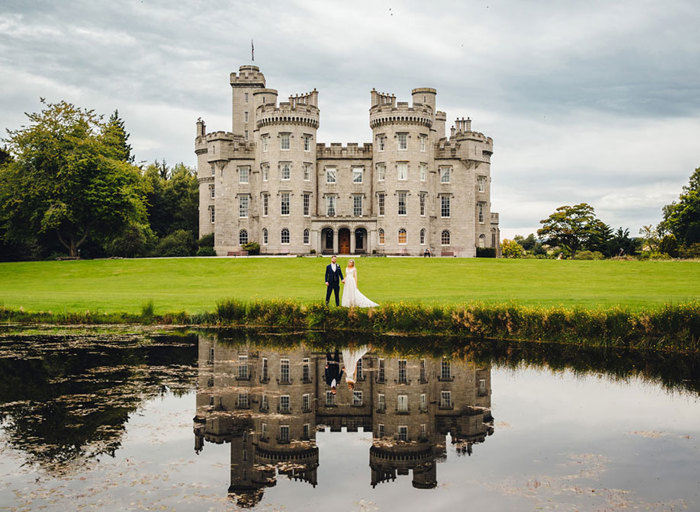 Bride and groom stand on the lawn outside Cluny Castle and their reflection is visible in the pond in front of them