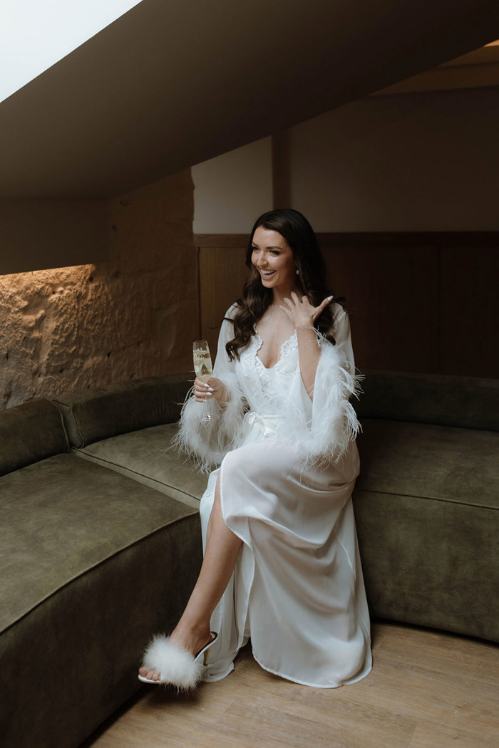 A bride wearing a silk and feather robe and a pair of white feather high heels holding a glass of champagne and laughing as she touches her hair