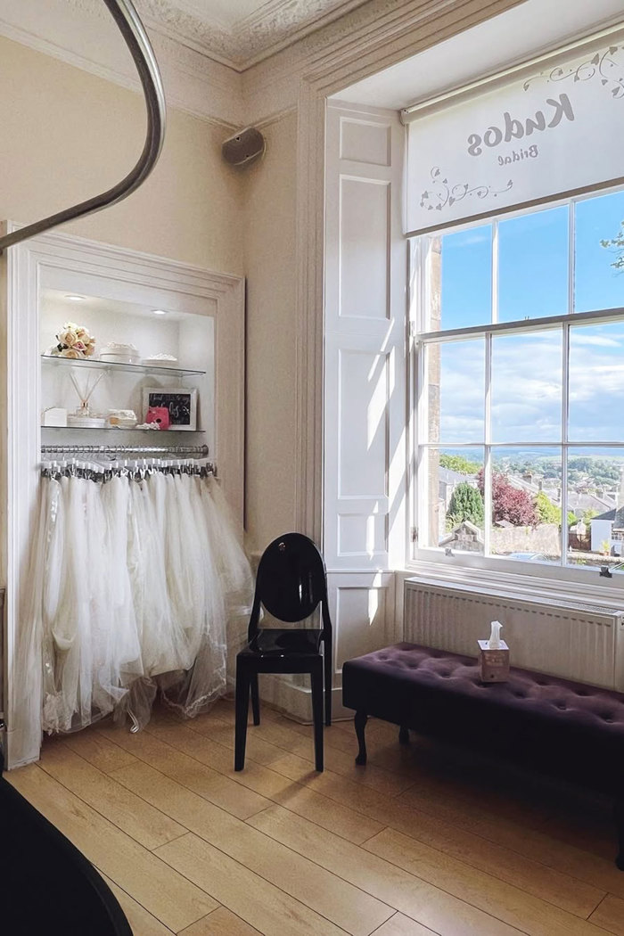 a view of inside a bridal boutique, with a look outside the window, a comfortable plush bench with tissues and a rack of wedding veils