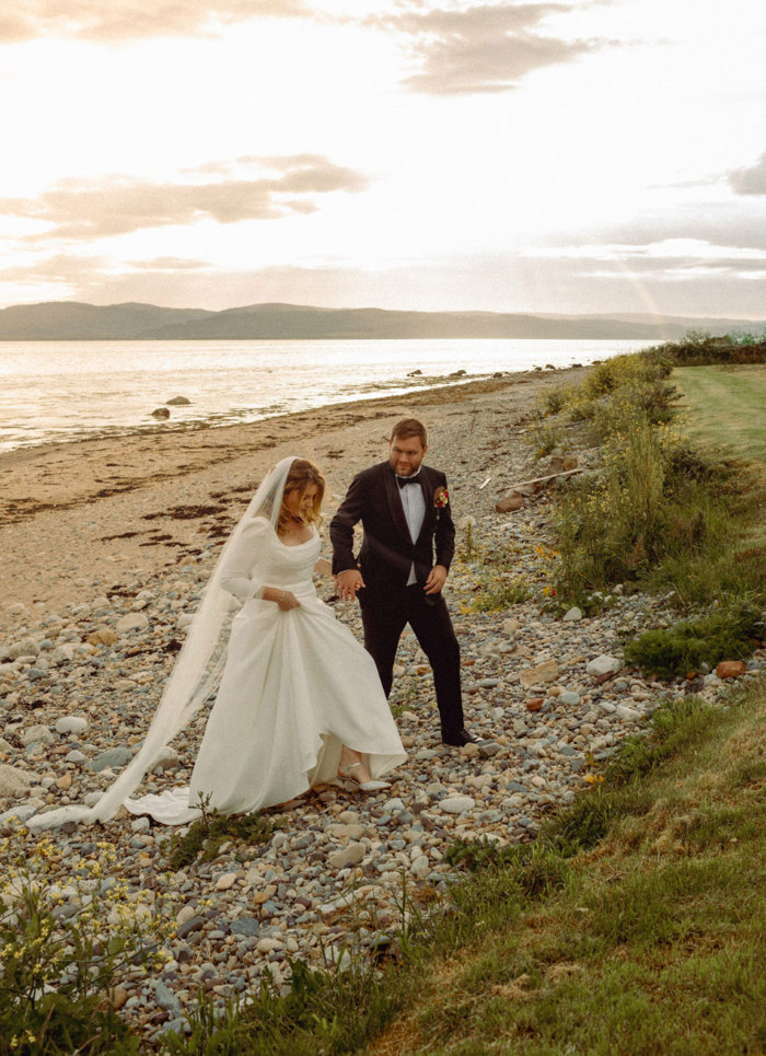 a bride and groom walking along a stony beach at sunset