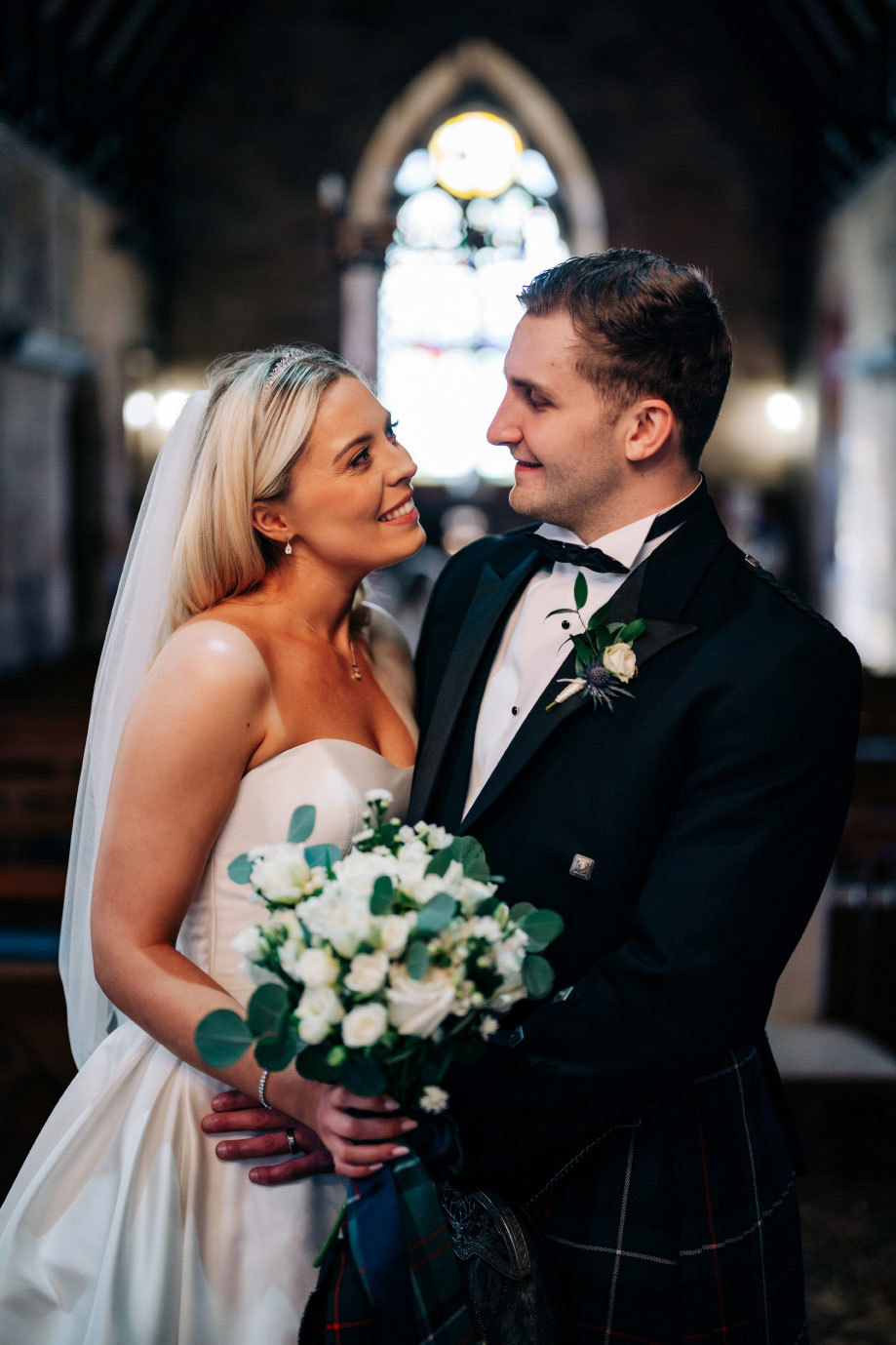 a bride and groom looking lovingly at one another. There is a blurred out stained glass window behind them