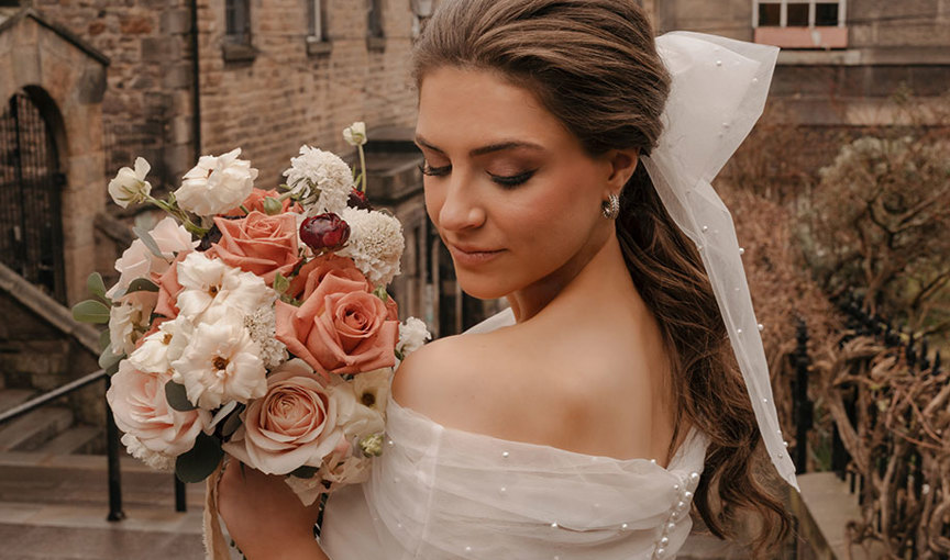 a person in a pearl scattered wedding dress wearing a large bow in hair holding a bouquet of pale pastel flowers