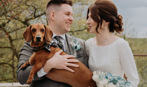  A man in a kilt holds a brown sausage dog who is wearing a bowtie as he stands next to a woman in a white dress