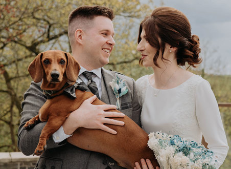  A man in a kilt holds a brown sausage dog who is wearing a bowtie as he stands next to a woman in a white dress