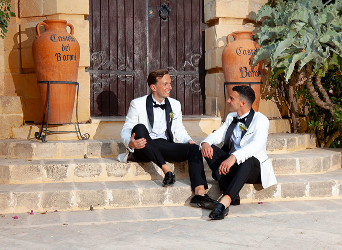 two grooms wearing smart white suit jackets and bow ties sitting on limestone steps in front of a large wooden door. There are two large terracotta urns either side of them reading 'Castello dei Baroni'