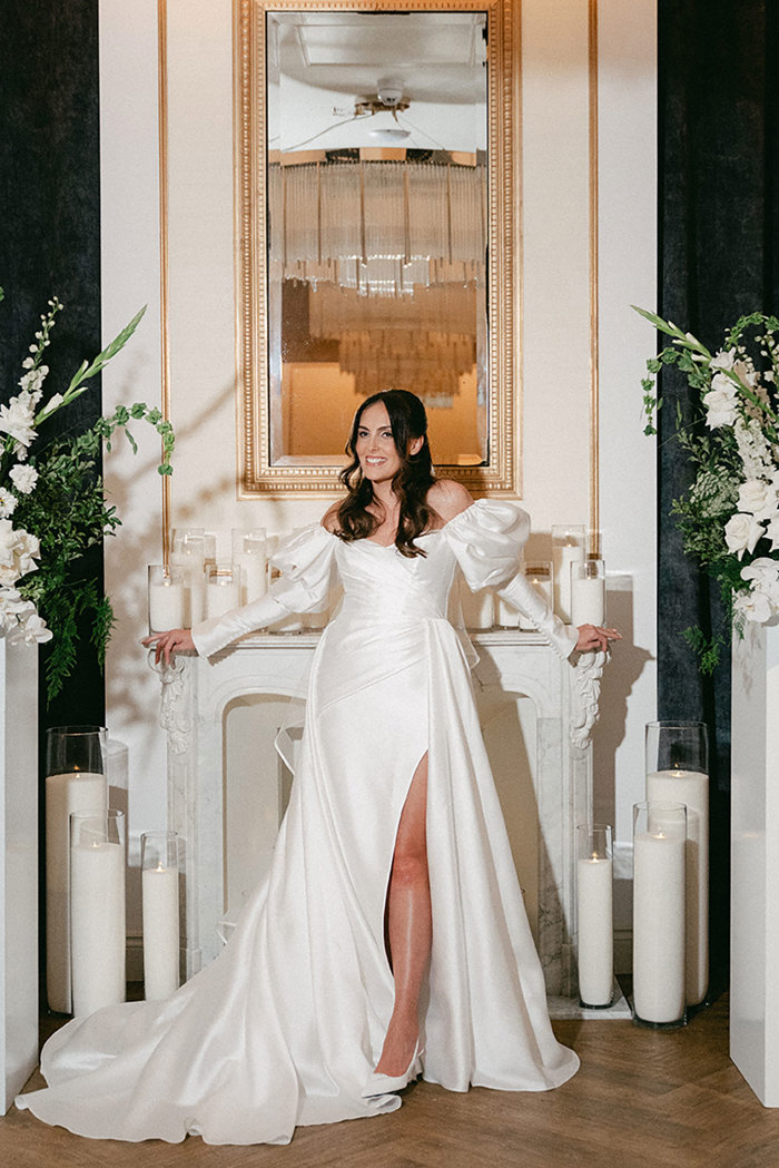brunette bride stands in front of white fireplace decorated with candles and flowers wears high leg slit wedding dress with puff sleeves