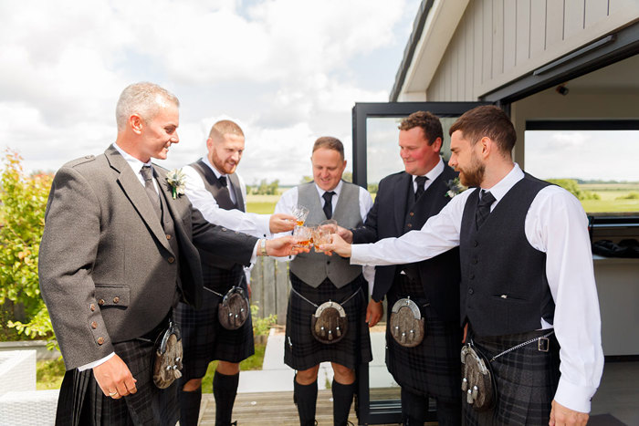 Five men in kilts clink glasses of whisky while standing in a circle 