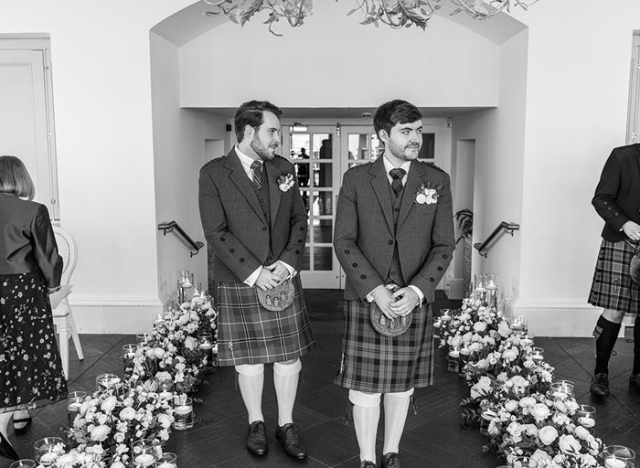 two grooms wearing kilts walking between two rows of white flower decorations on the floor