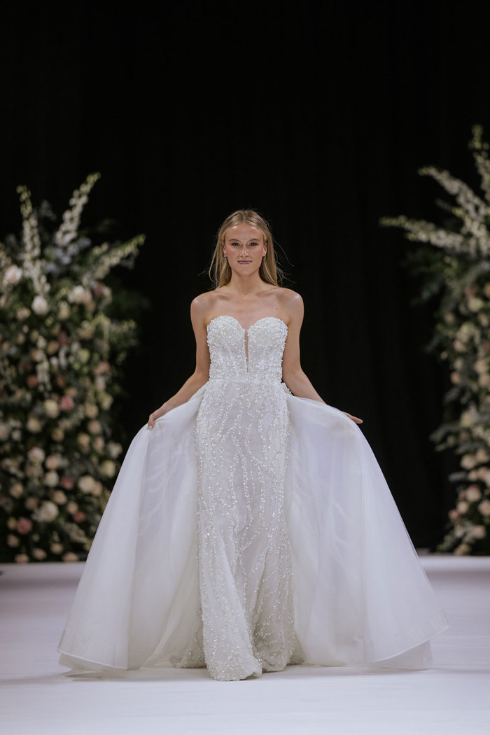 a model wearing a white strapless wedding dress on a catwalk. There are two tall flower displays in the background