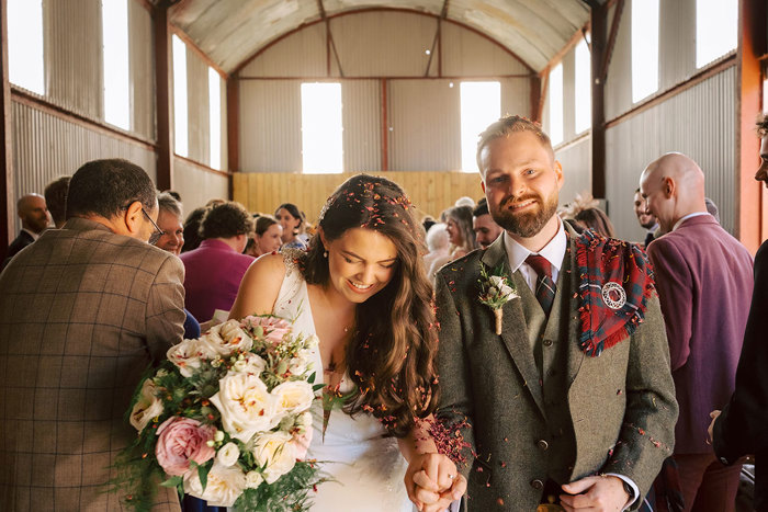 A bride and groom covered in confetti as they leave their ceremony with their guests behind them