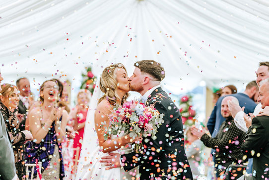 bride and groom kiss while being showered in multicoloured confetti as guests clap in background