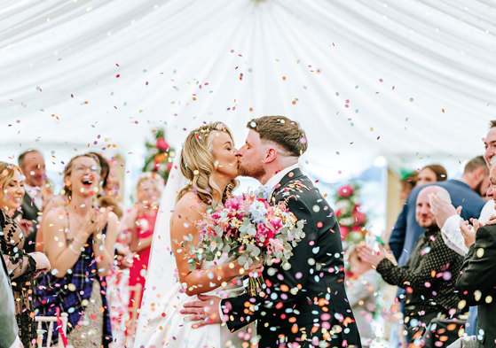 bride and groom kiss while being showered in multicoloured confetti as guests clap in background