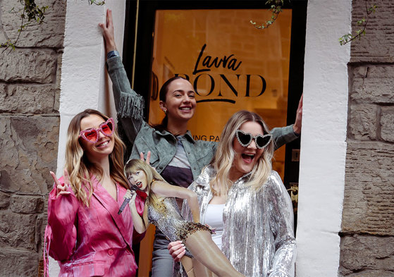 Three women stand in front of a glass door with the words 'Laura Bond' written on it, wearing heart shaped sunglasses and holding a Taylor Swift cardboard cutout