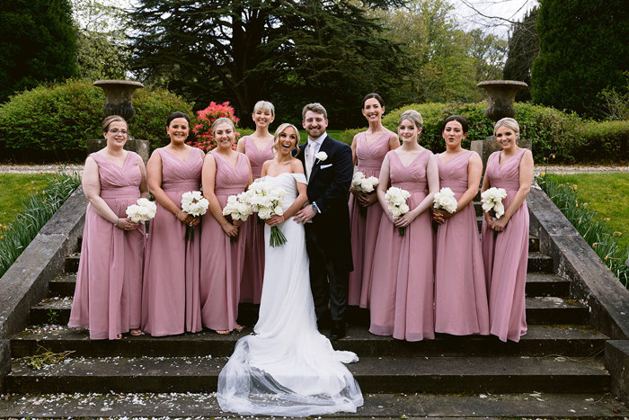 a bride and groom posing on a set of stairs at Mar Hall with eight bridesmaids wearing pink dresses.