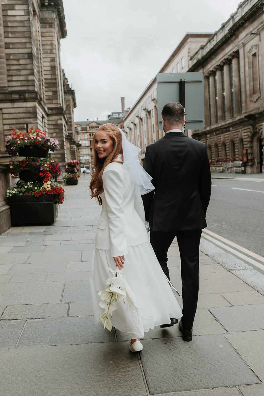 red haired bride turns back and looks at the camera as she walks hand in hand with groom through the street