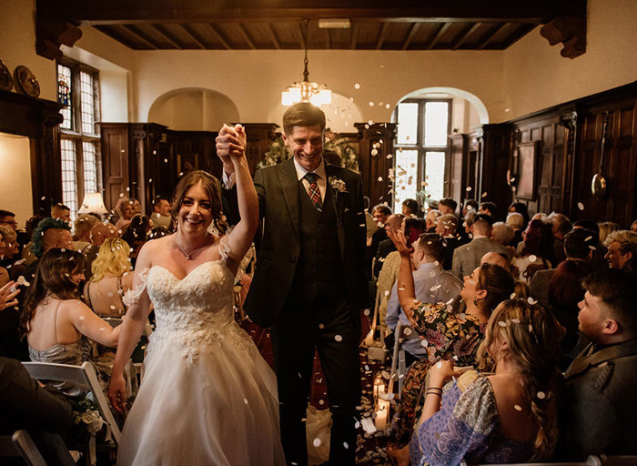 A bride and groom hold their clasped hands in the air as confetti is thrown over them