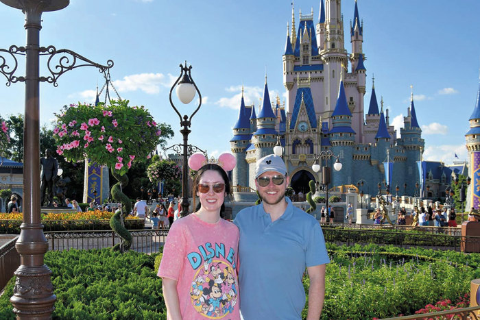 woman in pink Minnie Mouse ears and t-shirt with arm around man in blue Mickey Mouse cap and t-shirt in front of Disney Castle