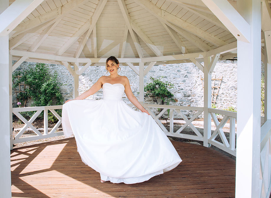 a bride in a white strapless gown holds her skirt out to both sides while standing in an outdoor gazebo