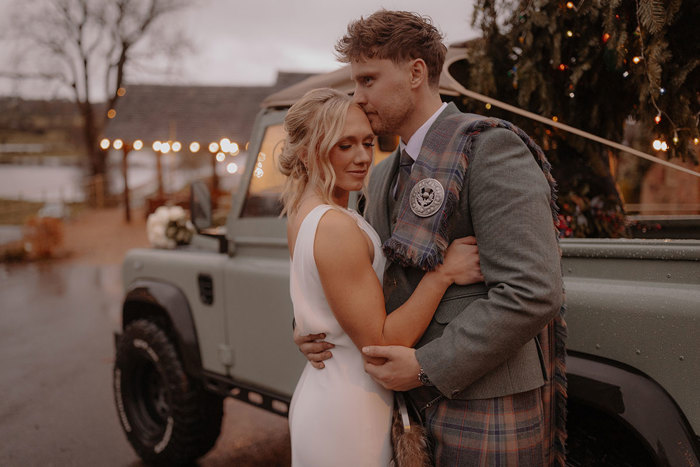 blonde woman in sleeveless white dress holds on to man in in green jacket and red, blue, green kilt with matching fly plaid, both stood in front of green truck with Christmas tree in the back 