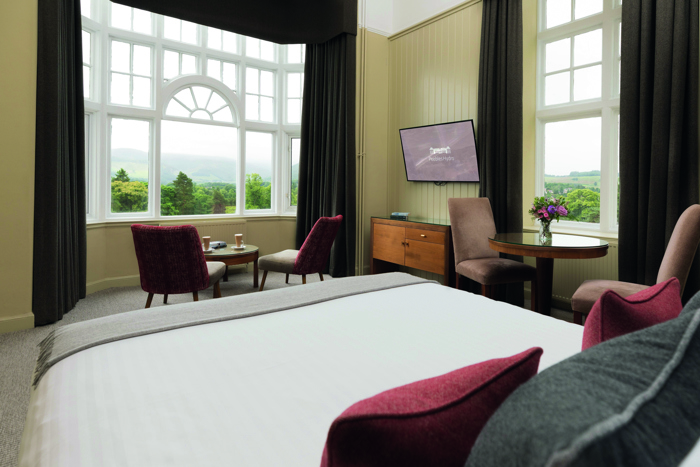 A hotel bedroom taken from the head of the bed looking at the rest of the room which includes two tables, four chairs and two windows with greenery outside