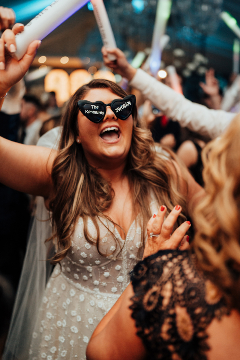 A bride wearing heart shaped sunglasses and holding a light up baton singing while on a dancefloor surrounded by people