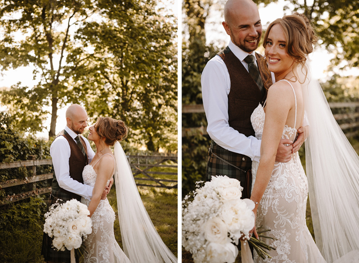 a bride and groom smile and pose next to a wooden gate and fence surrounded by verdant trees and hedges