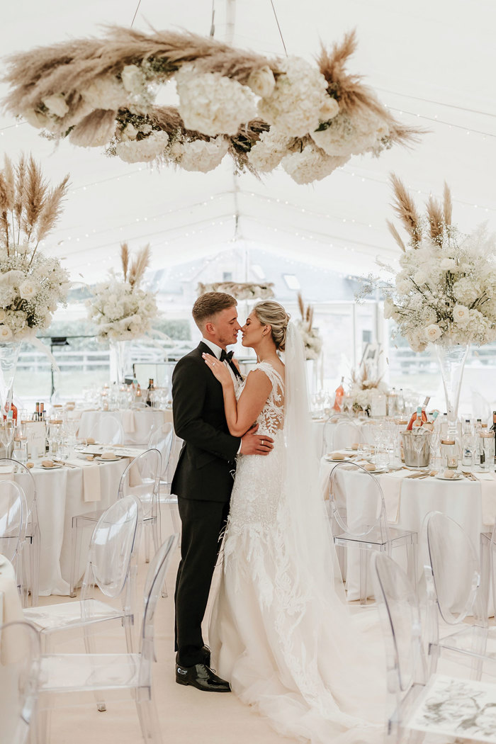 bride and groom lean in for a kiss while stood closely chest to chest in a white reception space filled with round tables and clear chairs