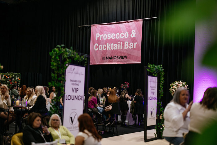 A group of people sitting at tables in front of a black curtain under a pink sign that reads "Prosecco and Cocktail Bar Manorview"