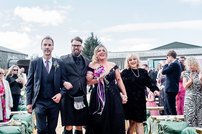 A Bride And Groom Wearing Black Walking Arm In Arm Outside With Two People, One Wearing A Black Short Dress And Another Wearing A Dark Grey Suit