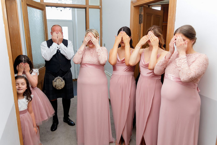 A group of women, girls and a man stand facing the camera with their hands over their eyes 