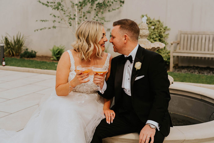 A bride and groom sitting on a stone bench holding drinks.