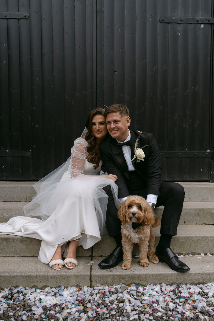 A bride and groom sitting on stairs with a dog outside the Engine Works.