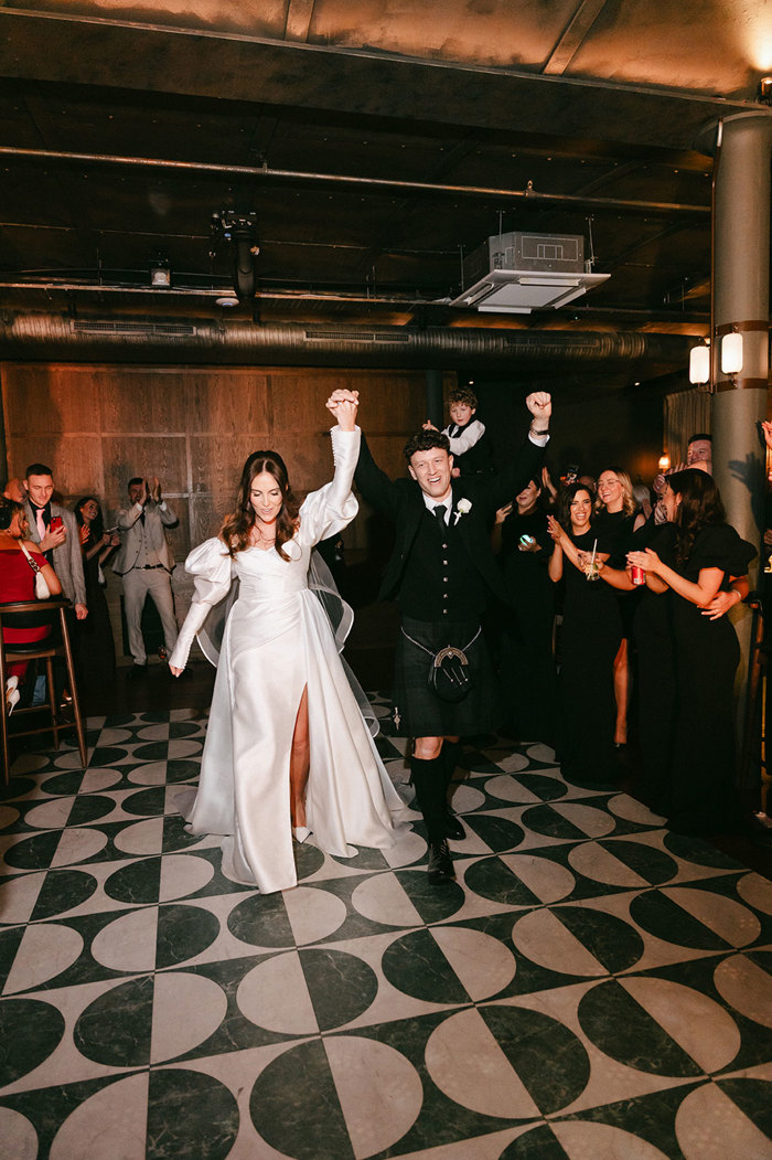 brunette bride in high leg slit wedding dress with puff sleeves and groom in full dark tartan kilt outfit walk with their hands intertwined above their heads