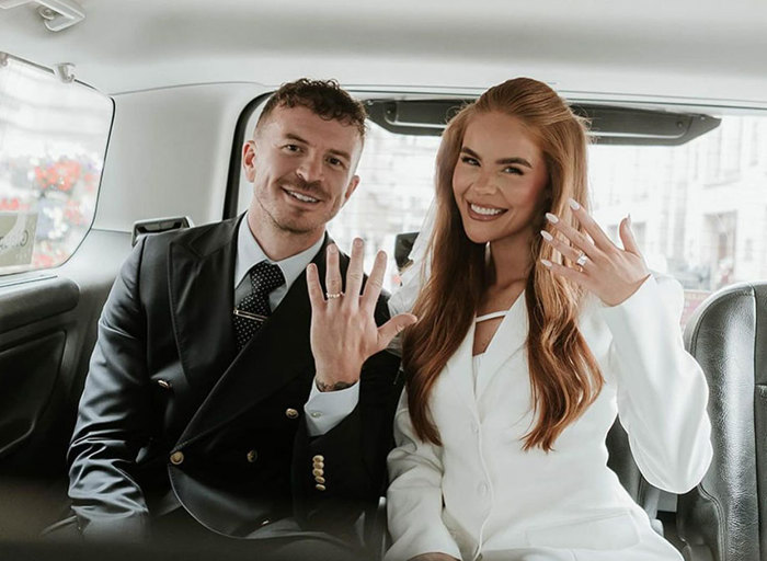 A man in a black suit and a woman in a white blazer hold up their left hands to the camera to show off their wedding rings while sitting in the back of a black cab taxi