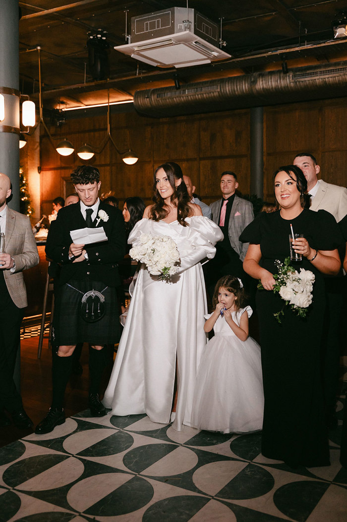 brunette bride in high leg slit wedding dress with long puff sleeves and groom in full dark tartan kilt outfit stand beside each other, bride smiling out at room as groom looks down at the piece of paper in his hands