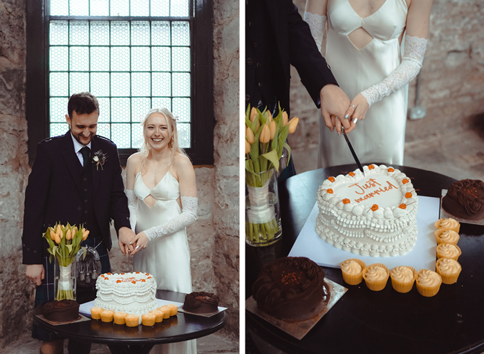 a bride and groom standing cutting a heart shaped wedding cake that's sitting on a round black table. Miniature cupcakes and a vase of yellow tulips also sit on the table on left. A heart shaped just married wedding cake with ornate swags and tails icing and cherries decorating it on right