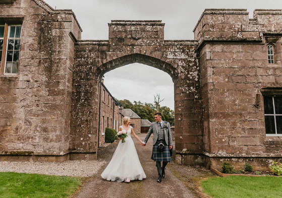 A bride in a long white dress and a groom in a blue and grey kilt hold hands and walk though a stone archway