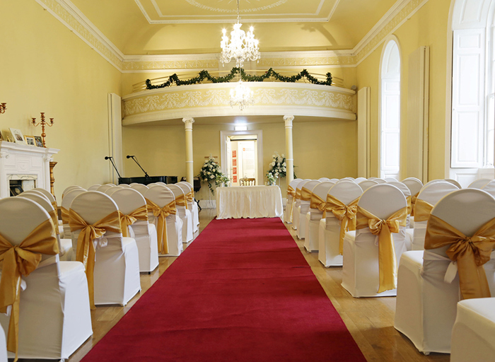 A venue with yellow walls set up for a wedding ceremony looking along a red aisle runner to a white table at the top of the aisle with white chairs which have gold ribbons tied on them along each side of the aisle