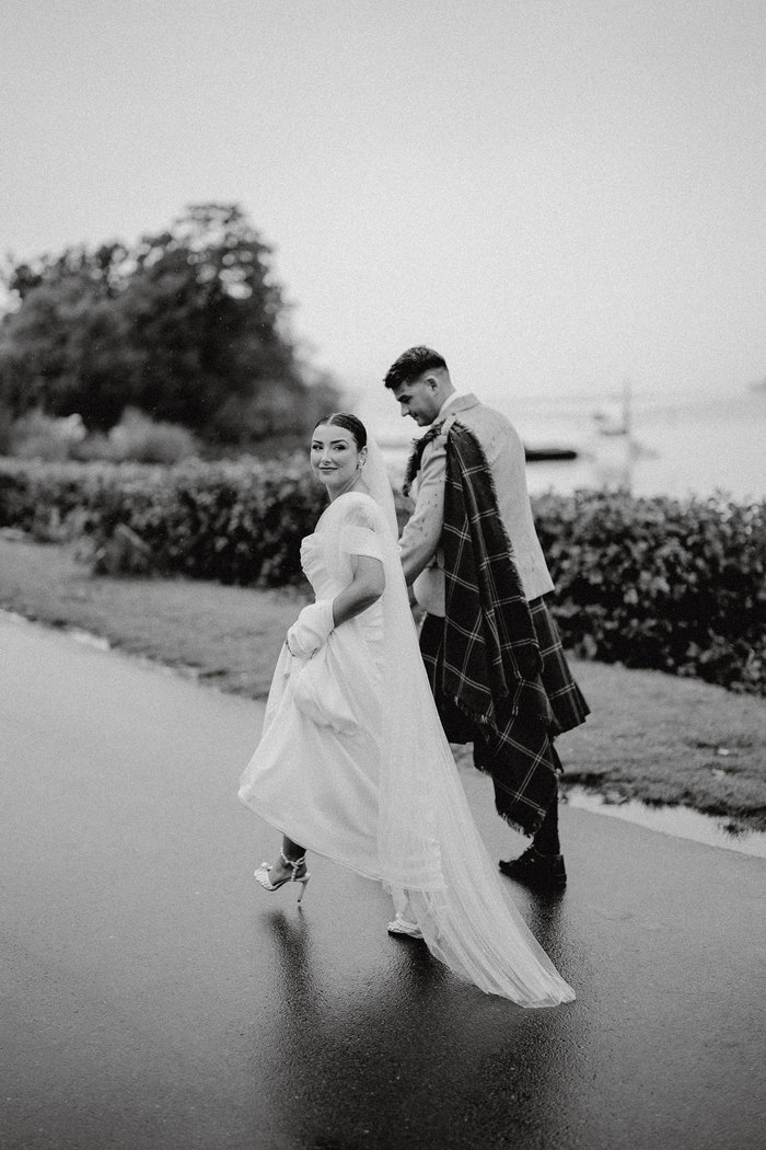 black and white photo of bride in strapless wedding dress with off the shoulder straps and a long trailing veil walks outdoors beside tall groom in kilt outfit