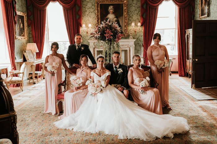 A group of well dressed people sitting on or standing behind a couch in a grand room decorated with paintings and flowers 