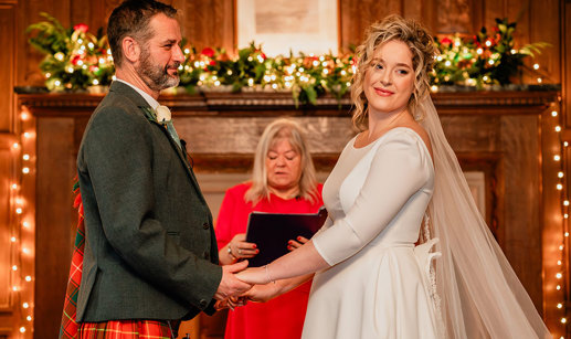 bride in white wedding dress and long veil and groom in red and green tartan kilt hold both hands out in front of them as a woman in front of them reads from a black folder