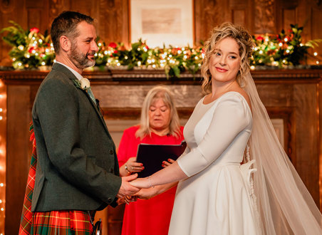 bride in white wedding dress and long veil and groom in red and green tartan kilt hold both hands out in front of them as a woman in front of them reads from a black folder