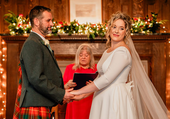 bride in white wedding dress and long veil and groom in red and green tartan kilt hold both hands out in front of them as a woman in front of them reads from a black folder